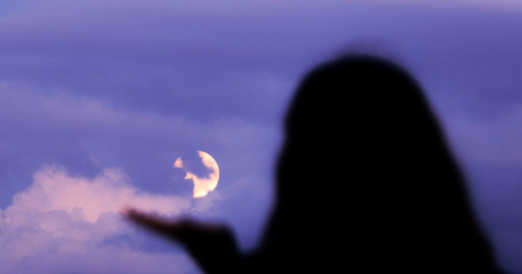 Silhouette of a woman reaching her open hand toward a glowing crescent moon in a dramatic purple twilight sky, surrounded by soft clouds.