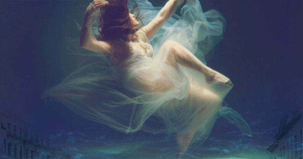 A red-haired woman in a flowing white tulle dress floating gracefully underwater, arms outstretched, with a blurred city skyline visible below her.