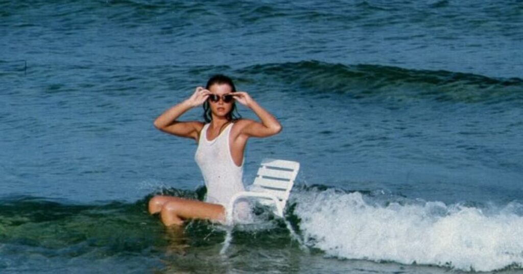 A woman in a white one-piece swimsuit and sunglasses sitting on a lounge chair in shallow ocean waves, holding her sunglasses as a wave crashes around her.
