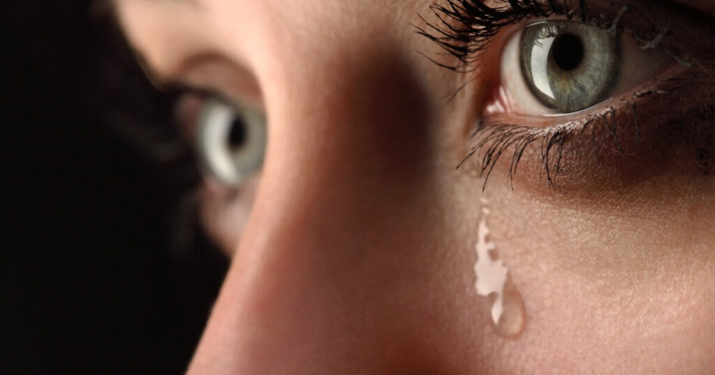 Extreme close-up of a woman's striking green eyes with a single tear rolling down her cheek against a dark background.