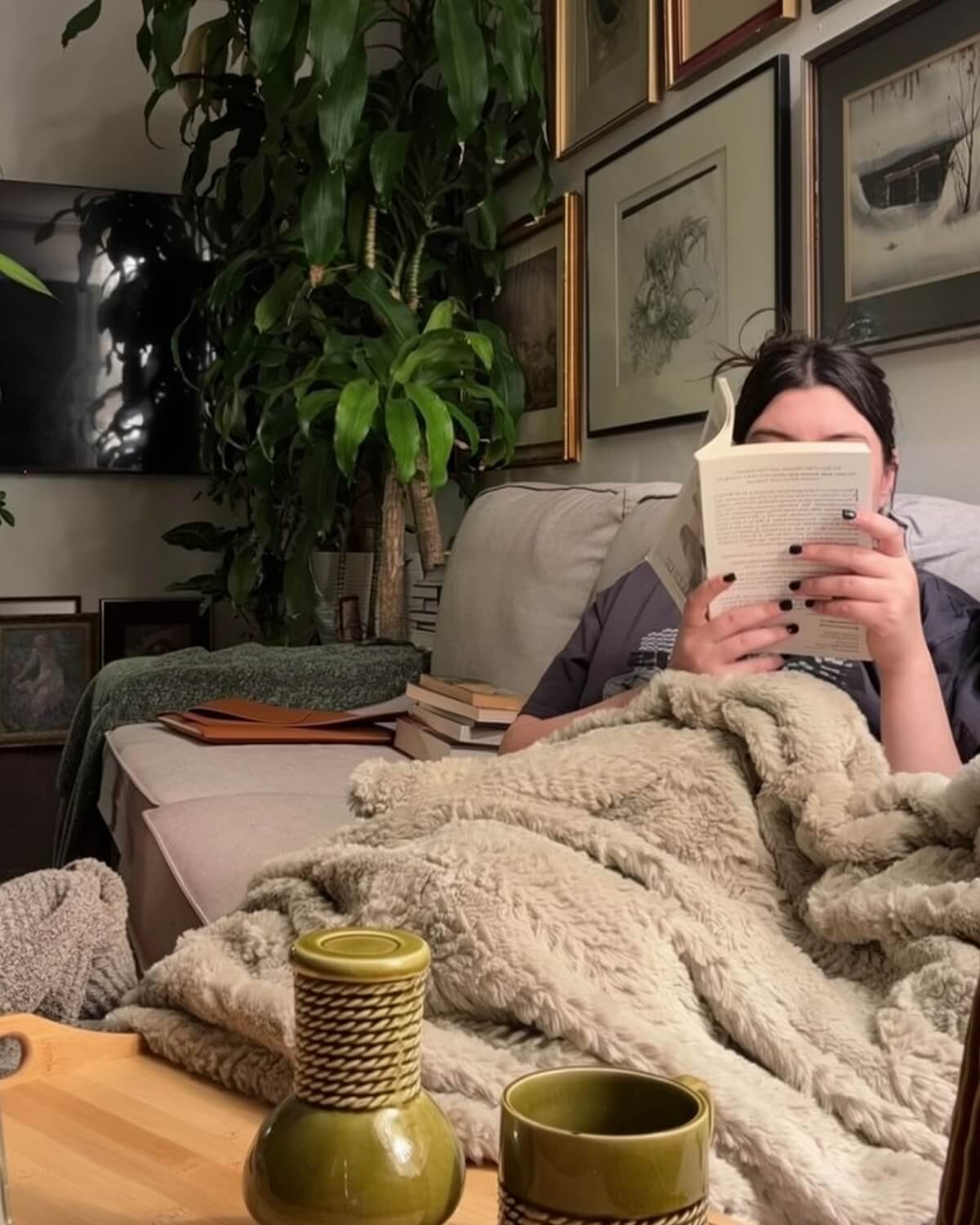 Woman reading a book on the sofa under a fluffy blanket with an olive green mug on the table, resting without guilt as part of a glow up