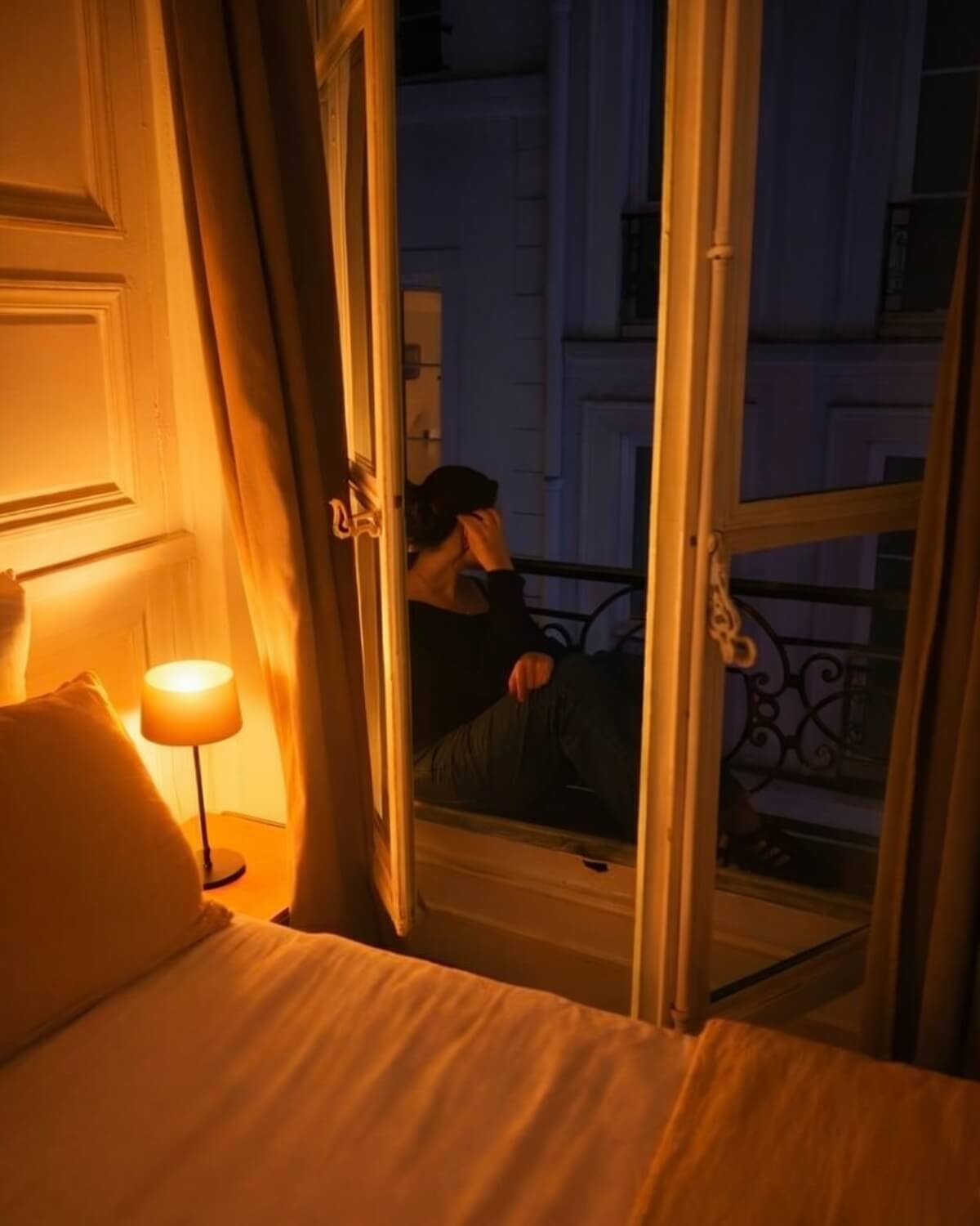 Woman sitting alone at an open French window at night with a warm bedside lamp glowing, practicing stillness as part of her evening ritual