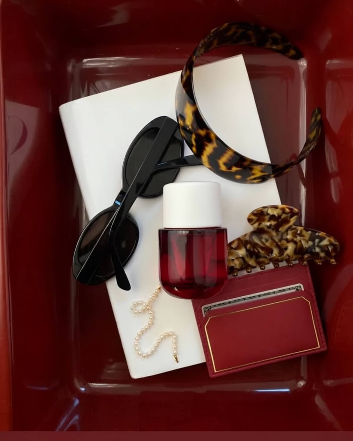 Flatlay in a burgundy tray with black sunglasses, a red perfume bottle, tortoiseshell hair accessories, pearls and a card holder, the signature details of your future self