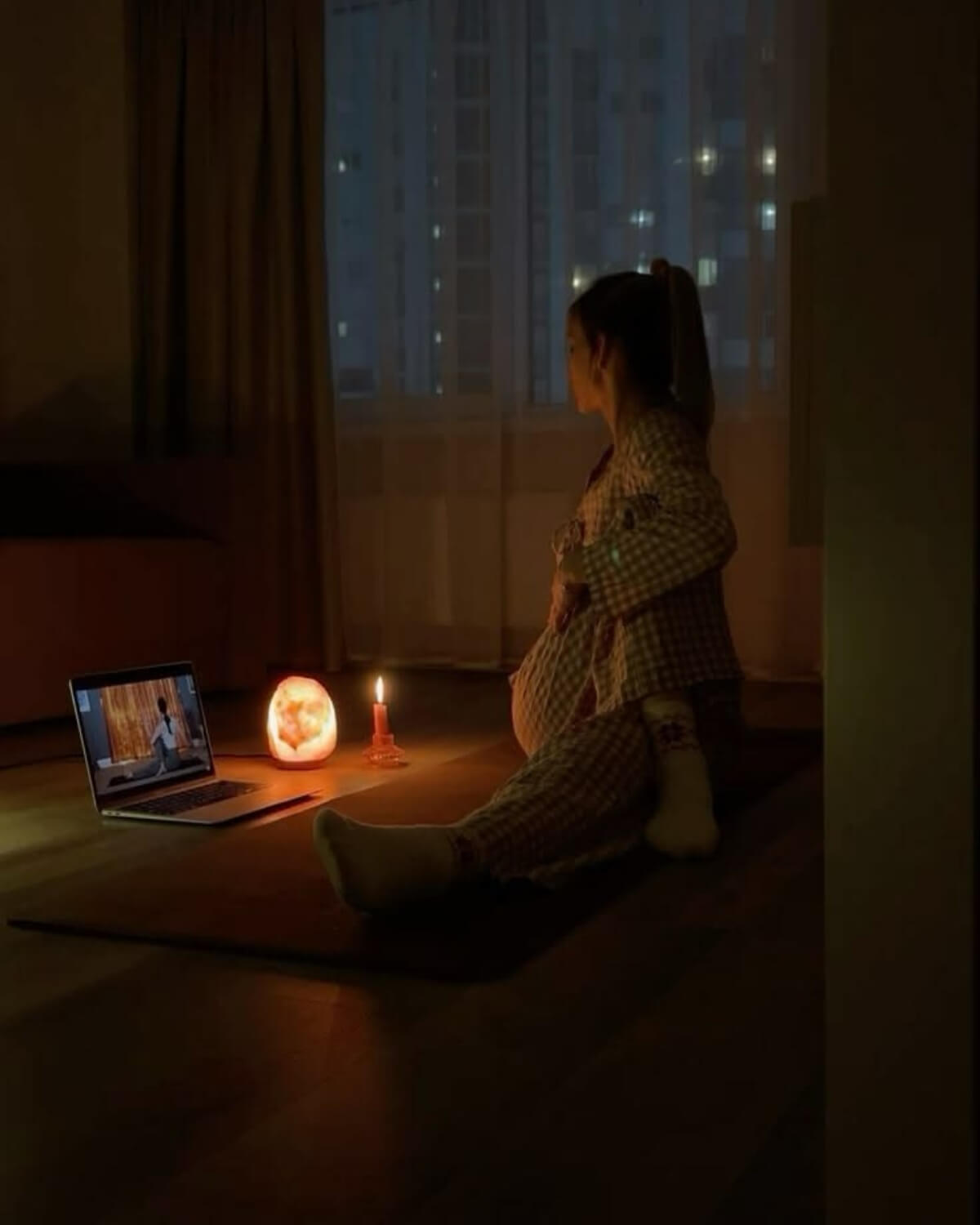 Woman in pajamas doing a gentle stretch on the floor by candlelight and a salt lamp at night, keeping her evening ritual on a low-energy night