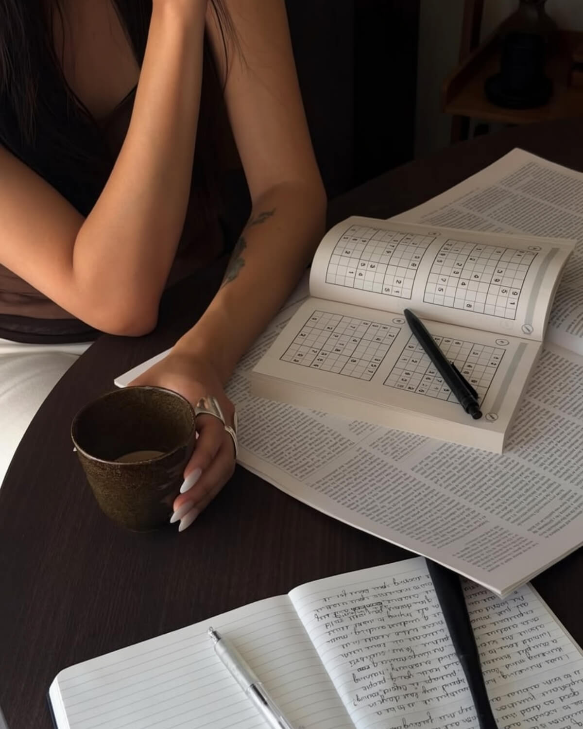 Woman at a dark wooden table with coffee, a sudoku book, a newspaper and a handwritten notebook, building the habit of becoming dangerously educated