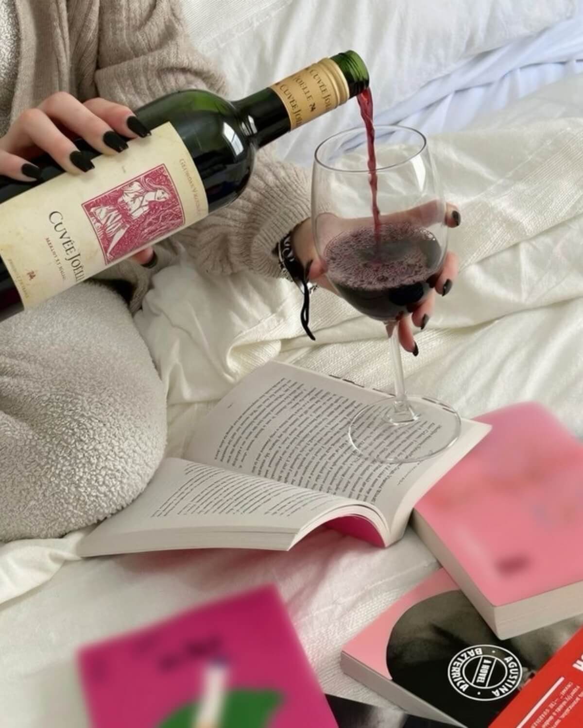 Woman in a cream sweater pouring red wine on a white bed surrounded by open books, living as her future self in an ordinary evening