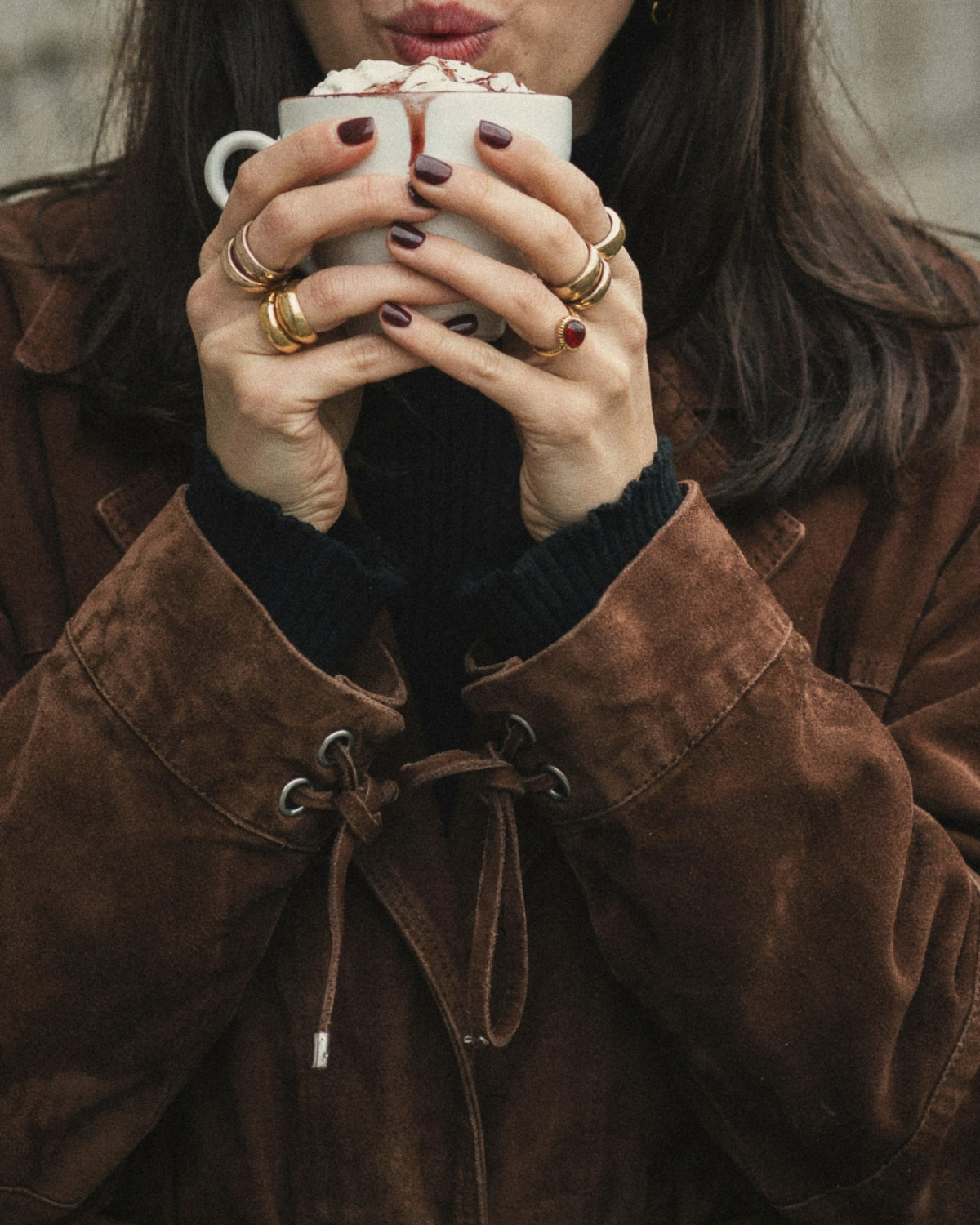 Woman in a brown suede jacket with burgundy nails and gold rings holding a cup of hot chocolate, calm and confident in an everyday moment