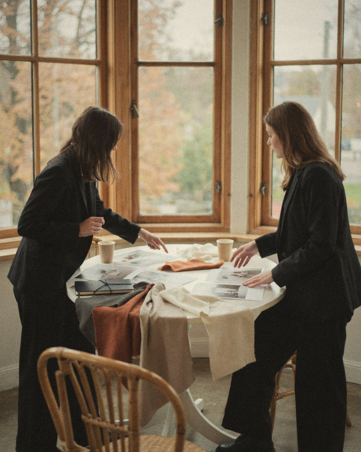 Two women in black blazers leaning over a round table with fabric swatches and prints by an autumn window, a calm first meeting