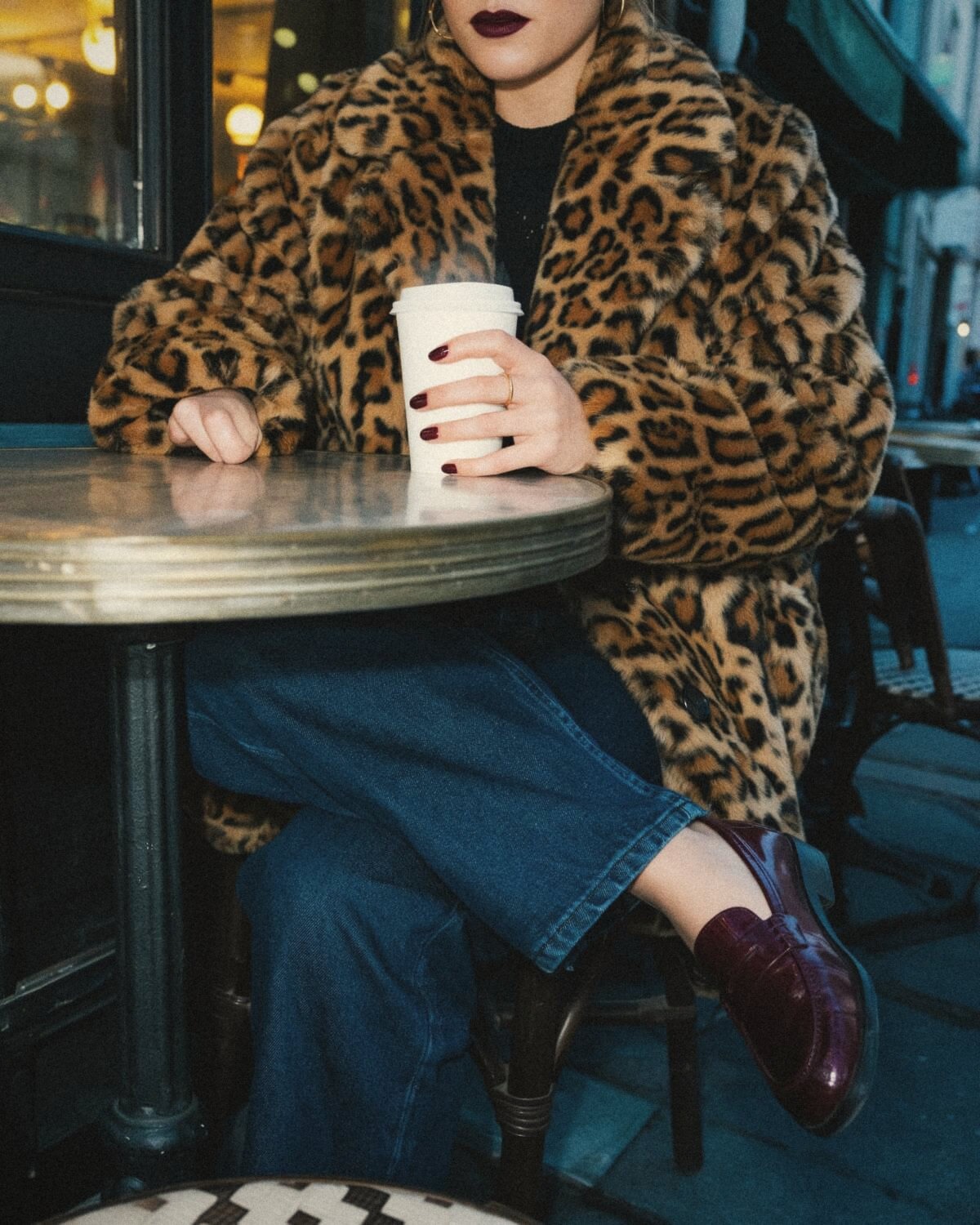 Woman in a leopard print coat, black knit, jeans and burgundy loafers holding a coffee at a café table, a casual dark feminine outfit