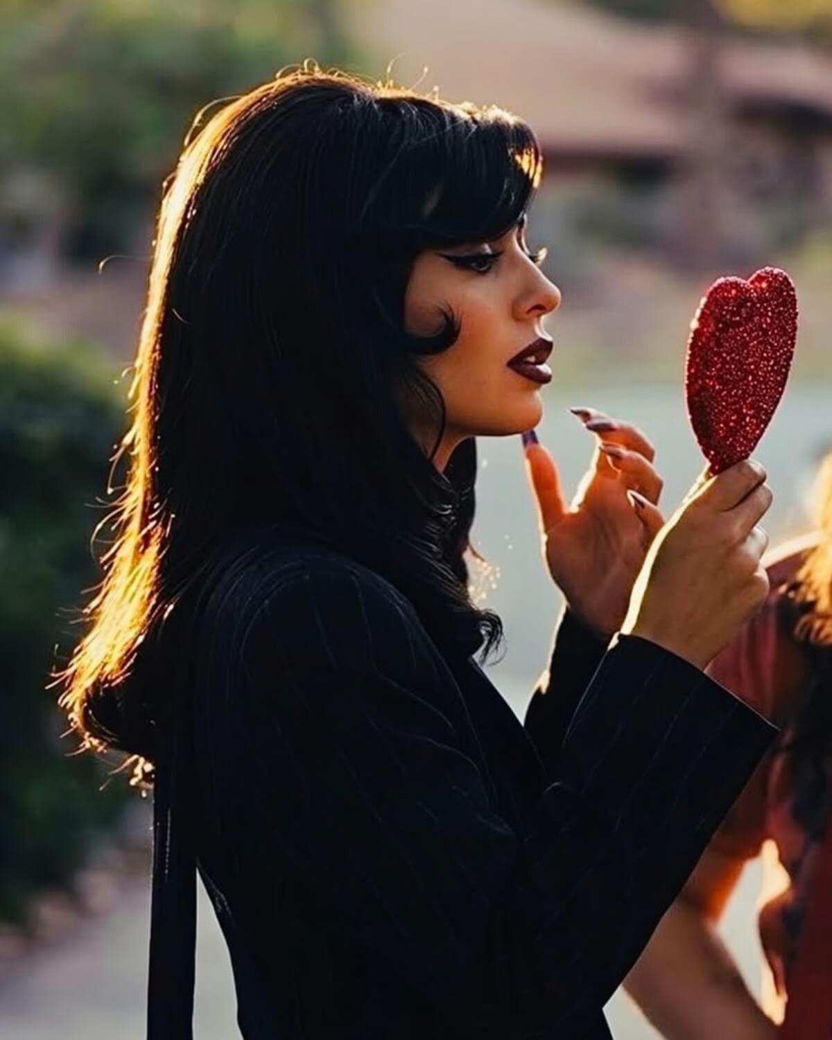 Woman with black hair, winged eyeliner and dark plum lipstick checking a red glitter heart mirror in golden light, dressing for herself