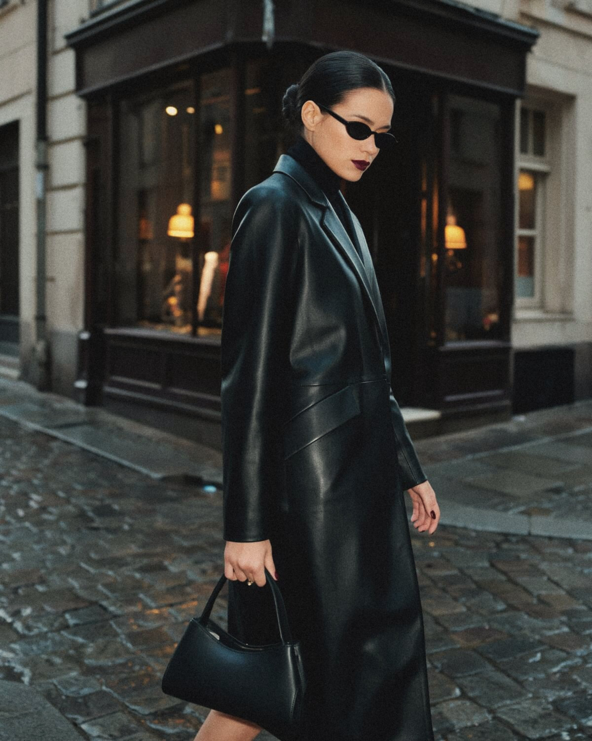 Woman with a sleek low bun, small black sunglasses and dark lipstick walking on a wet cobblestone street in a long black leather coat, an everyday dark feminine outfit