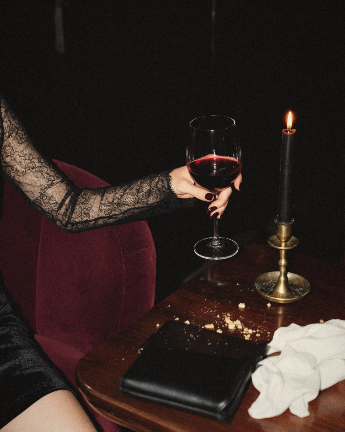 Arm in a sheer black lace sleeve holding a glass of red wine beside a burgundy velvet chair and a black candle, dark feminine fabrics in one frame