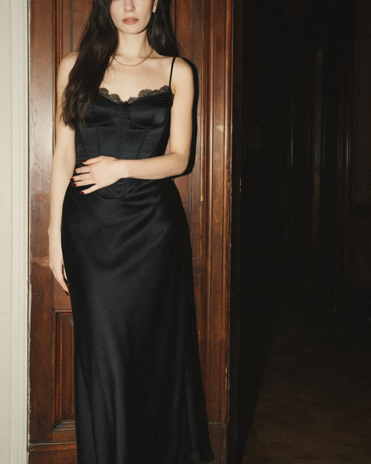 Woman leaning against a dark wooden door in a black satin corset top with lace trim and a long fluid satin skirt, a structured and flowing dark feminine silhouette