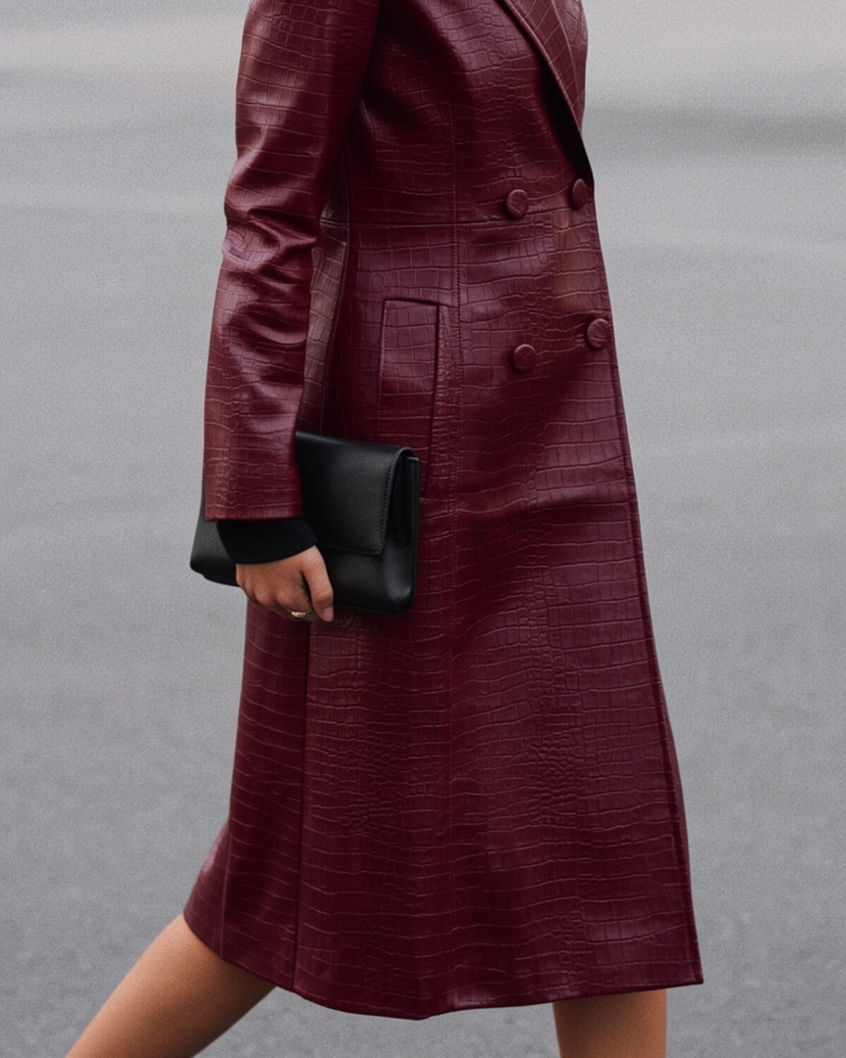 Woman in a long burgundy croc-embossed leather coat walking slowly with a black clutch, showing calm dark feminine presence