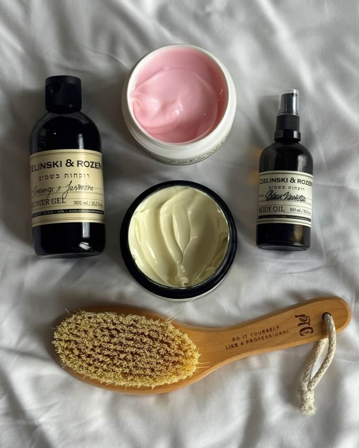Flatlay of a shower gel, body oil, two jars of body cream and a wooden dry brush on white sheets, a simple body care routine for a 30 day glow up challenge