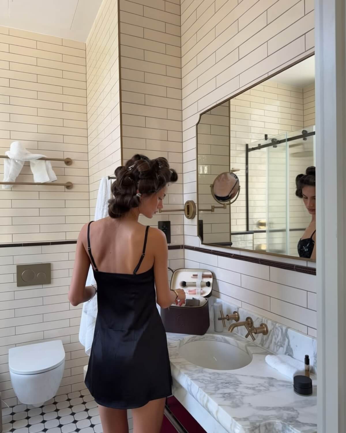 Woman in a black satin slip dress getting ready in a marble hotel bathroom with hair rollers, acting like her future self before she feels ready