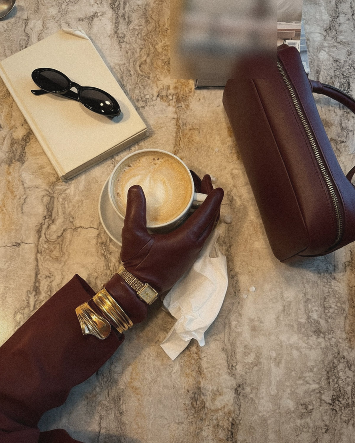 Hand in a burgundy leather glove with gold bangles holding a cappuccino on a marble table next to a burgundy bag, lifestyle branding in everyday details