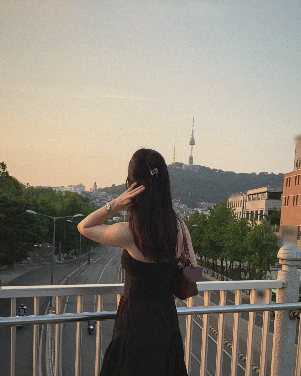 Woman with long dark hair on a Seoul footbridge at sunset looking toward N Seoul Tower, a new city before her first networking event