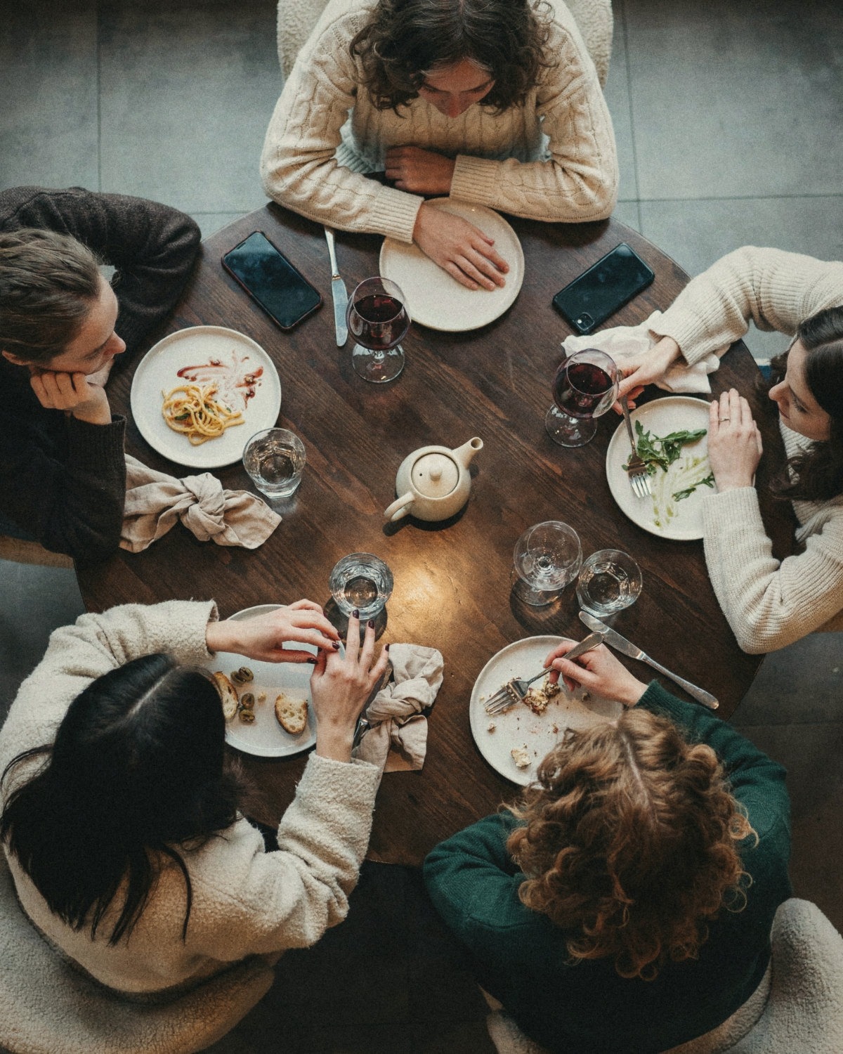Overhead view of five women sharing dinner and wine around a round wooden table, a social connection that grew into friendship