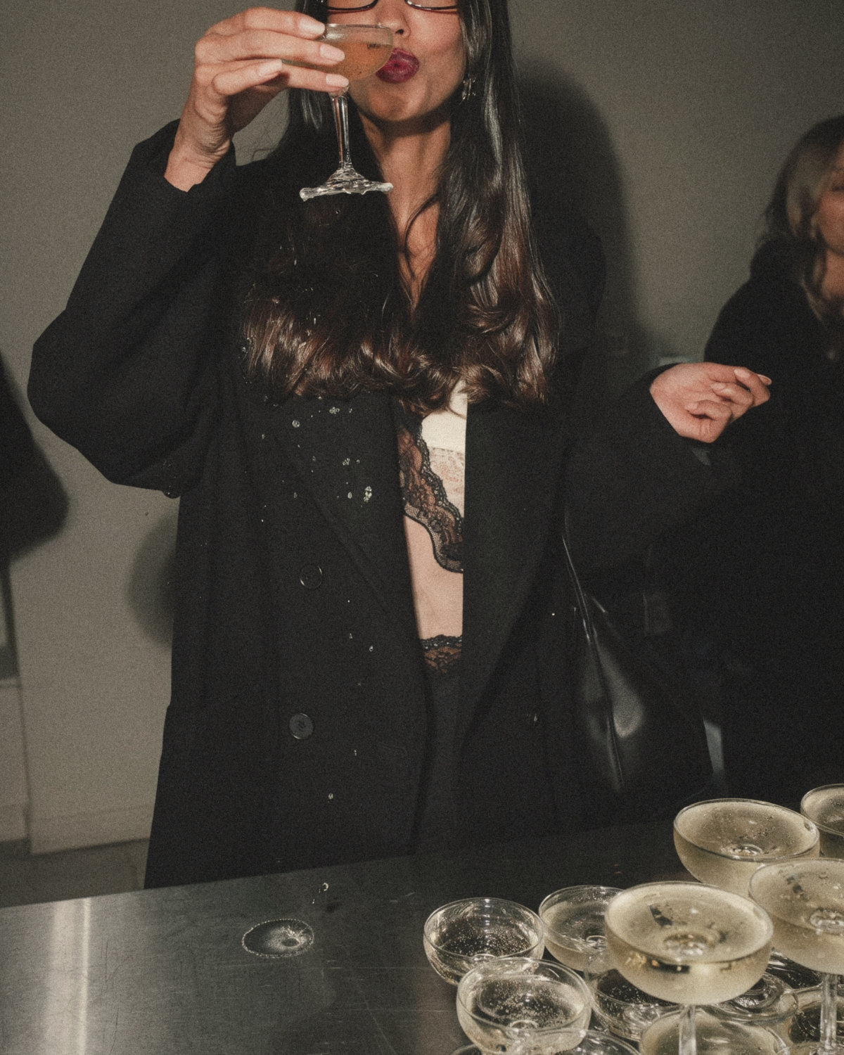 Woman in a black coat and lace top raising a coupe of champagne at an evening event, enjoying social networking her own way