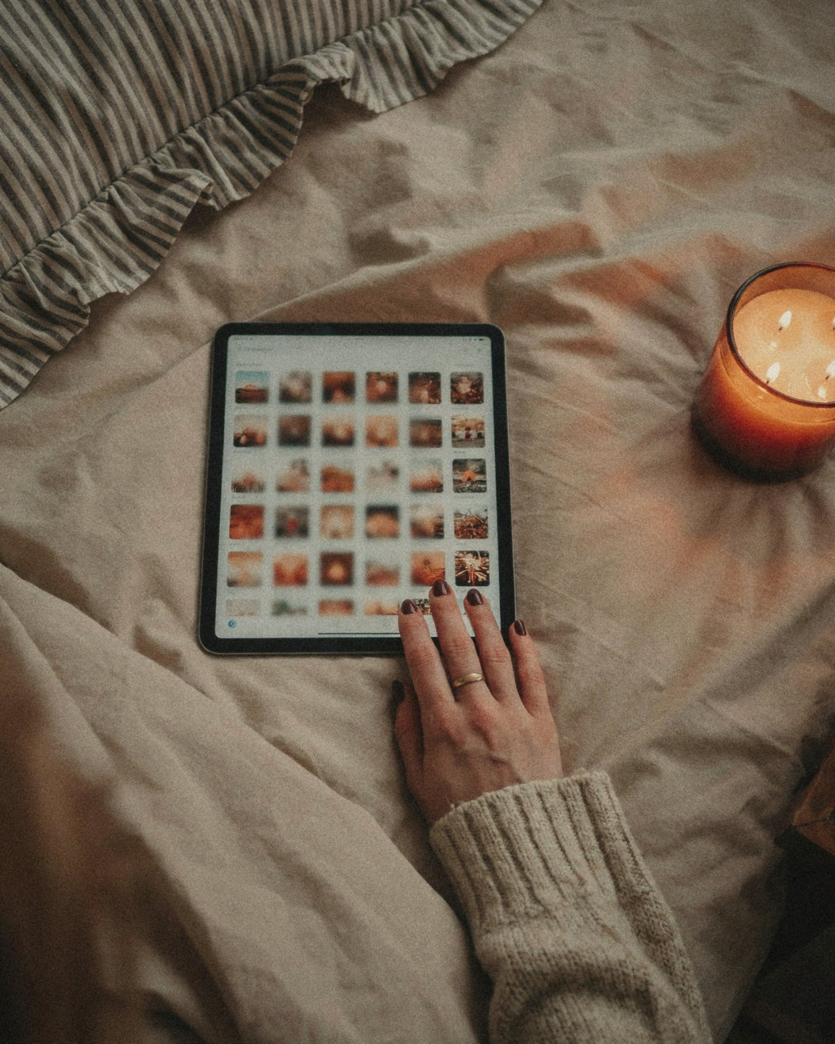 Hand with burgundy nails scrolling a grid of photos on a tablet on a bed by candlelight, auditing an online presence at night