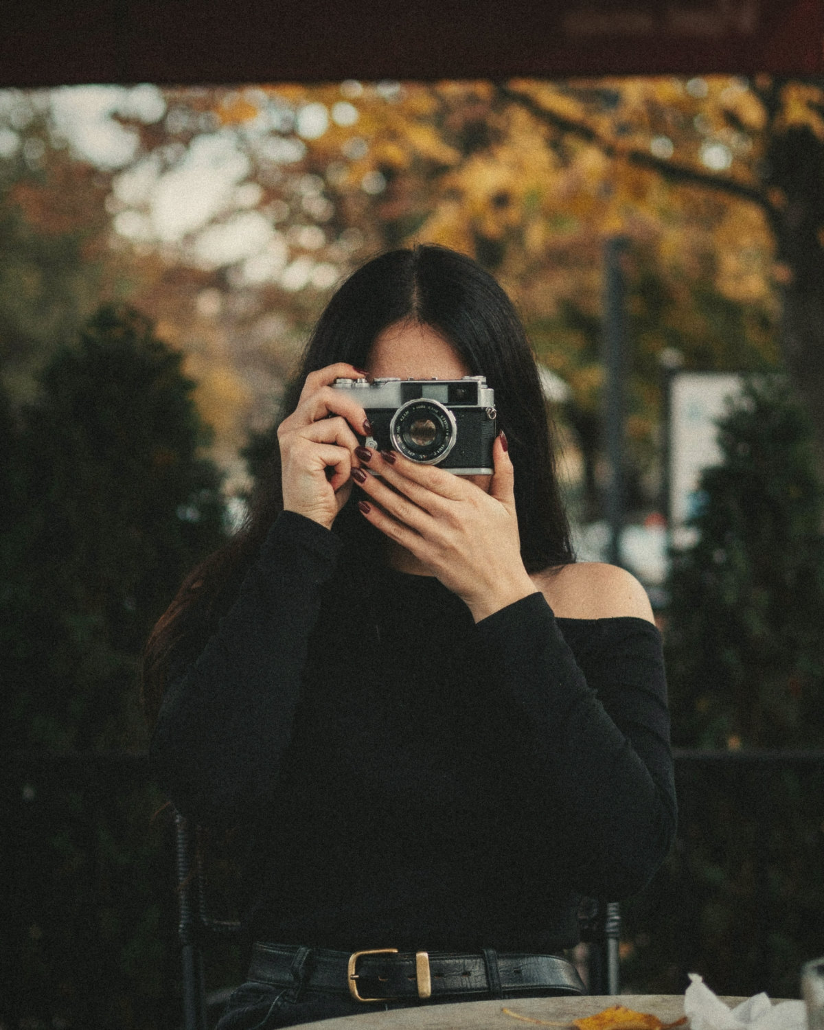 Woman with long black hair and burgundy nails holding a vintage film camera on an autumn café terrace, capturing moments for her online presence