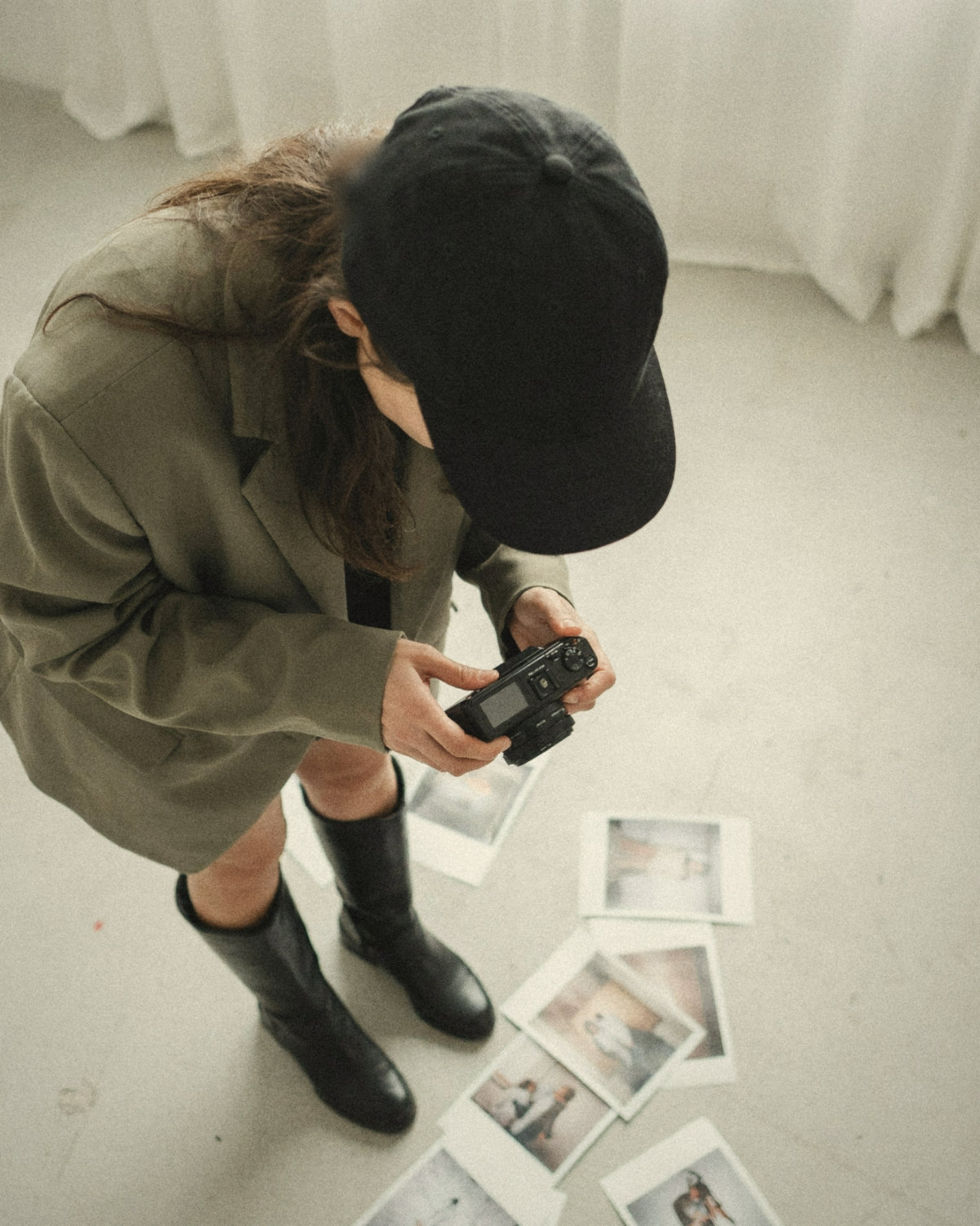 Woman in a black cap and olive blazer reviewing shots on a camera above printed photos on the floor, choosing what her online presence should say