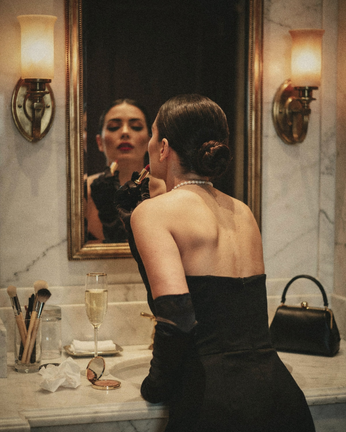 Woman in a black strapless dress, opera gloves and pearls applying red lipstick in a gold mirror, her real self matching her online presence