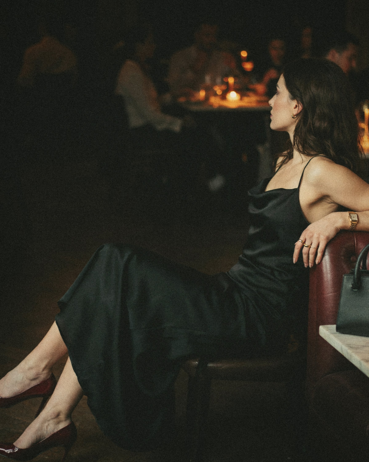 Woman in a black satin slip dress and burgundy heels sitting in a candlelit restaurant looking away, a mysterious and intentional online presence