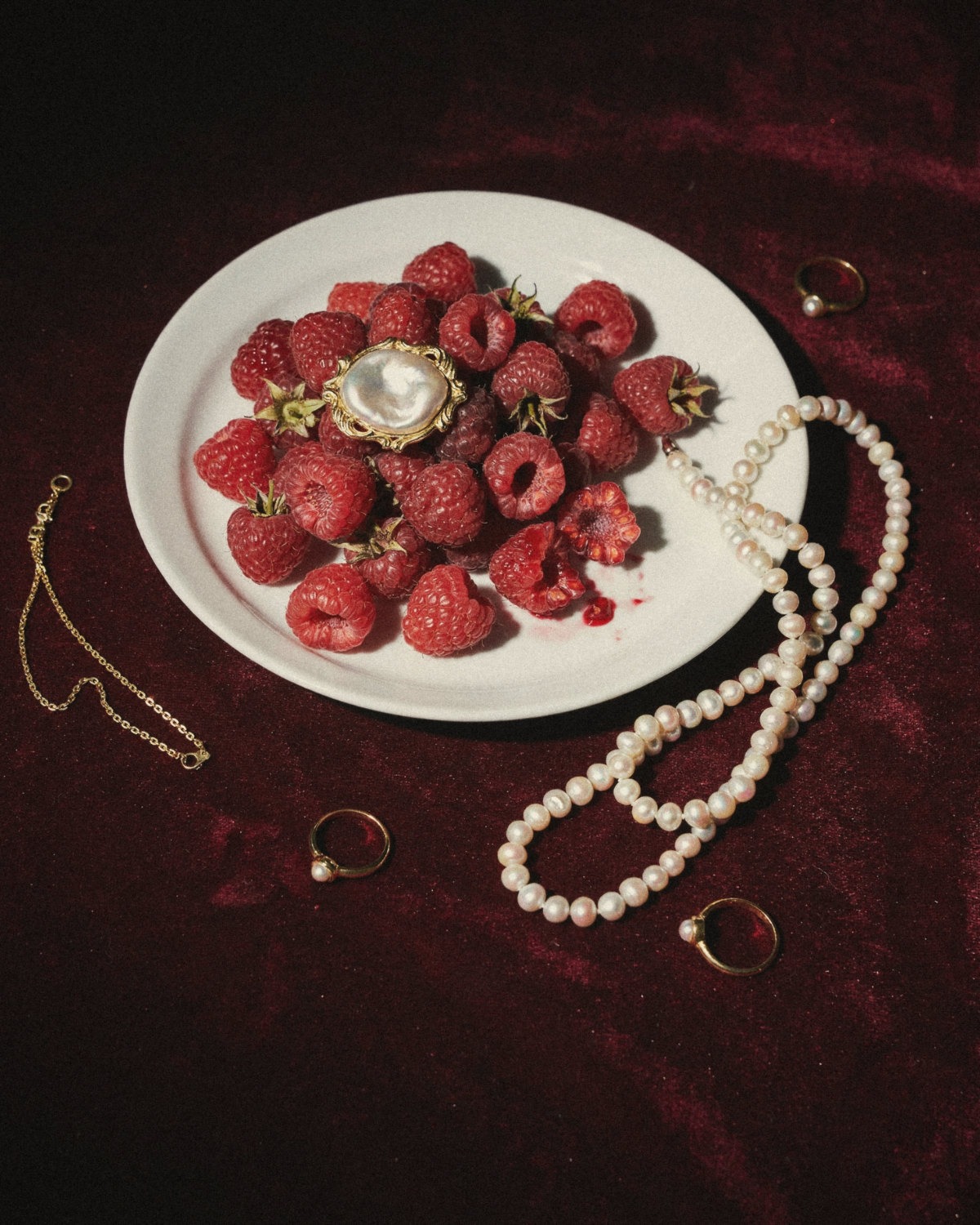 Plate of raspberries with pearl necklaces and gold rings on deep burgundy velvet, a signature color palette for a curated online presence