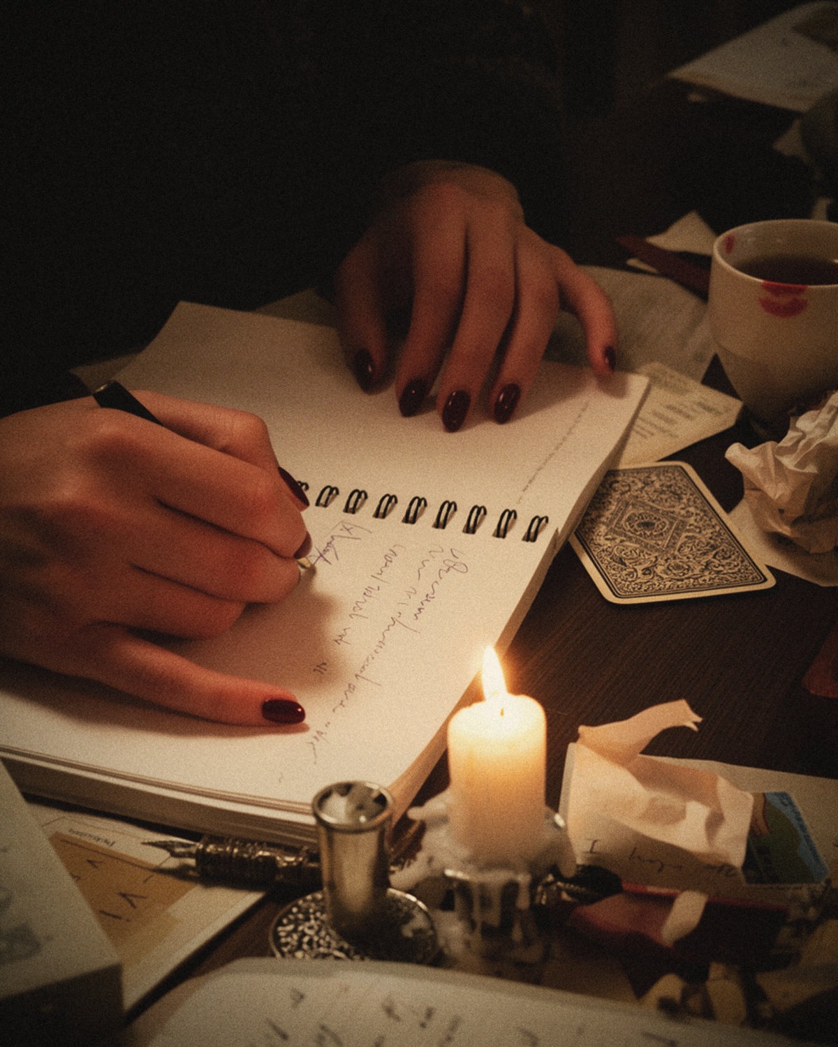 Hands with dark burgundy nails writing in a notebook by candlelight next to a tarot card, defining a personal brand identity