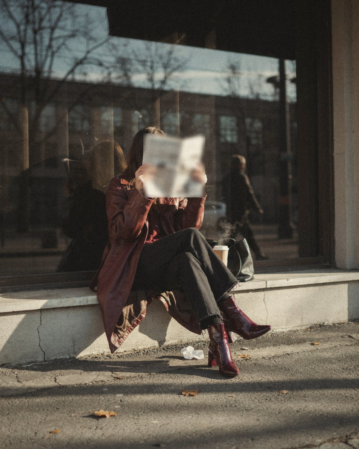 Woman in a long burgundy leather coat and burgundy patent boots reading a newspaper on a city ledge, a signature color as part of her personal brand