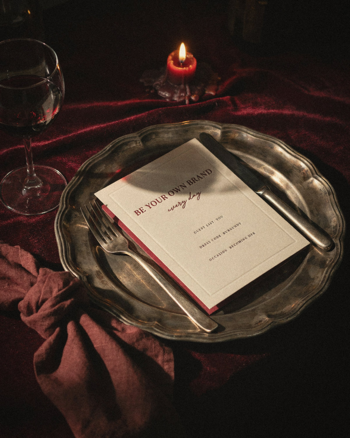 Candlelit dinner table with a silver plate, red wine and a cream card reading be your own brand every day, personal branding inspiration